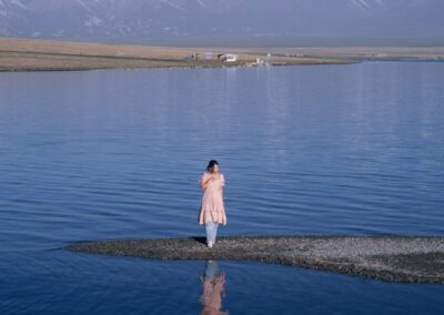 Woman on land strip in lake with snow-capped mountains.
